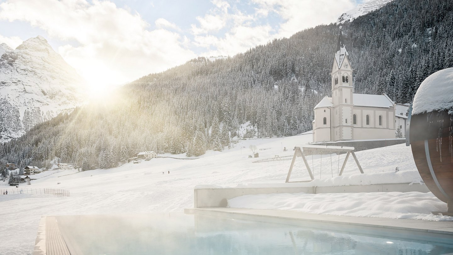 Beheizter Außenpool des Hotel Bella Vista bei Sonnenuntergang mit Sonnenstrahlen, die durch die Bäume scheinen und den Dampf über dem Wasser in der schneebedeckten Landschaft Südtirols beleuchten.