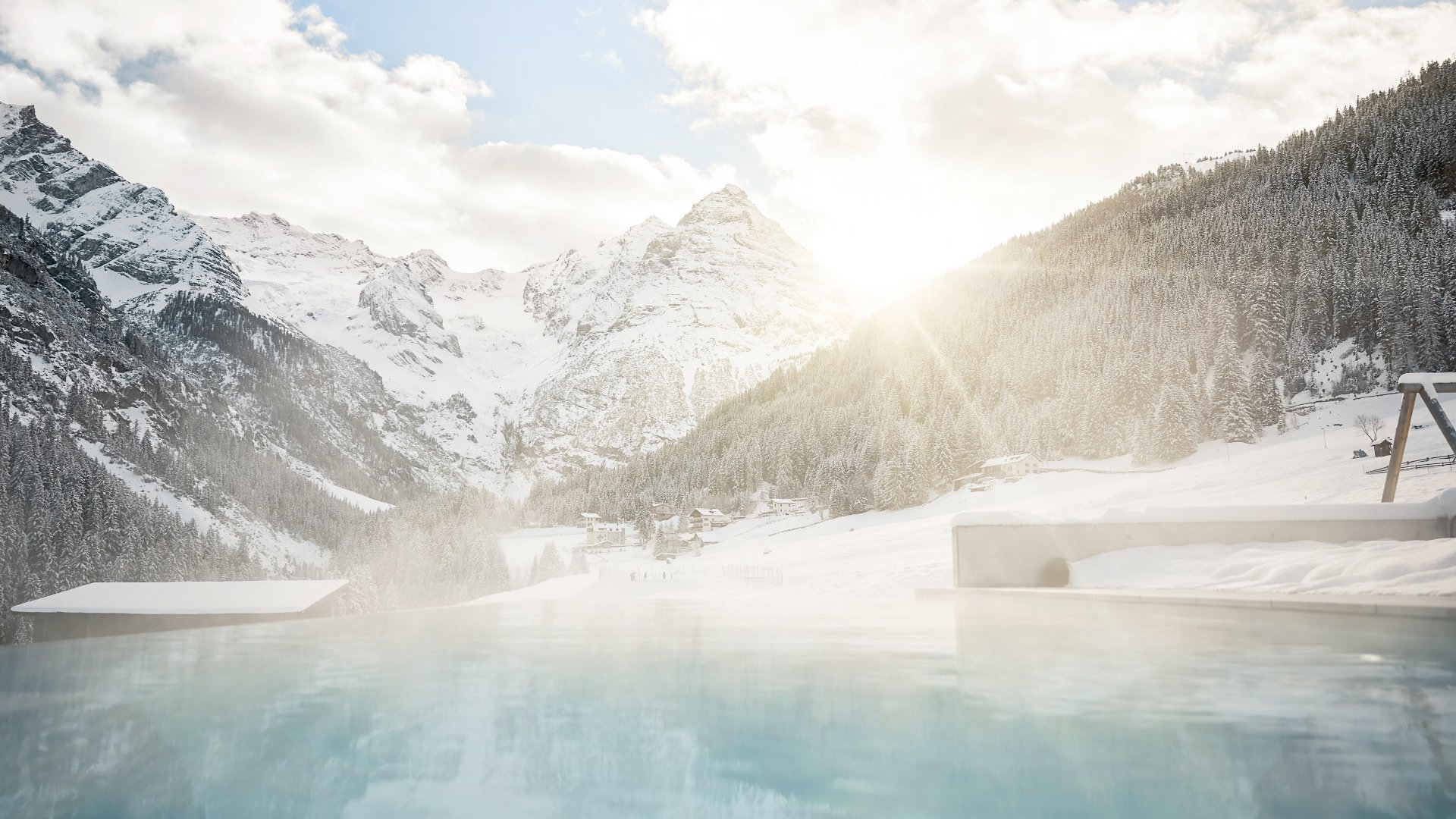 Winterliches Schwimmvergnügen im beheizten Außenpool des Hotel Bella Vista mit Blick auf verschneite Bäume und die Bergkulisse im Sonnenuntergang.