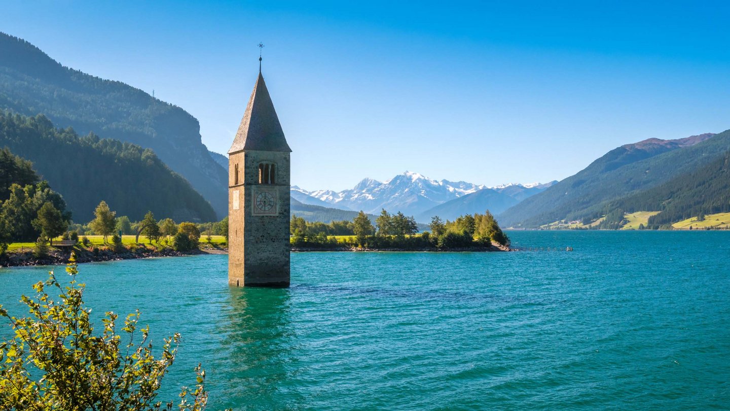 The submerged bell tower of Graun emerges from the clear blue waters of Lake Reschen, surrounded by vibrant greenery and the majestic mountain scenery of South Tyrol.