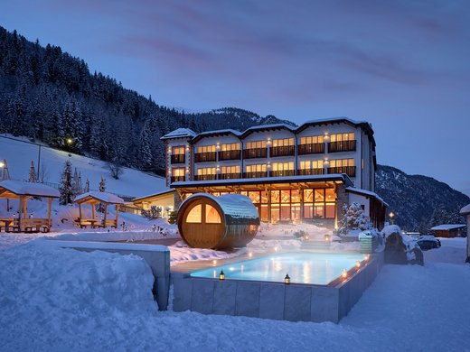 Abendansicht des Hotel Bella Vista in Südtirol mit beleuchteten Fenstern und einem dampfenden Außenpool, umgeben von einer schneebedeckten Berglandschaft.